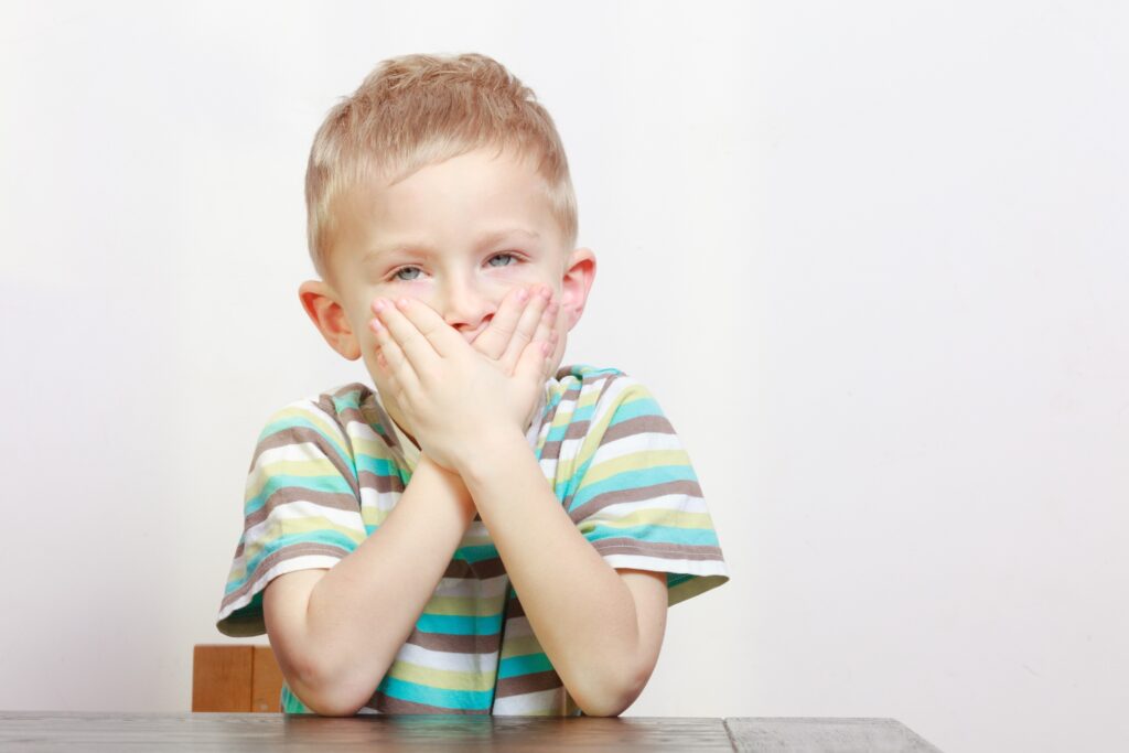 Young blond boy covering his mouth with both hands.